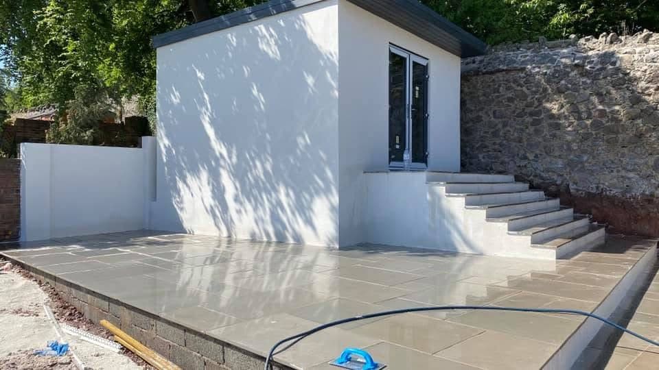 Modern white garden studio with glass doors, steps, and a large grey stone patio.